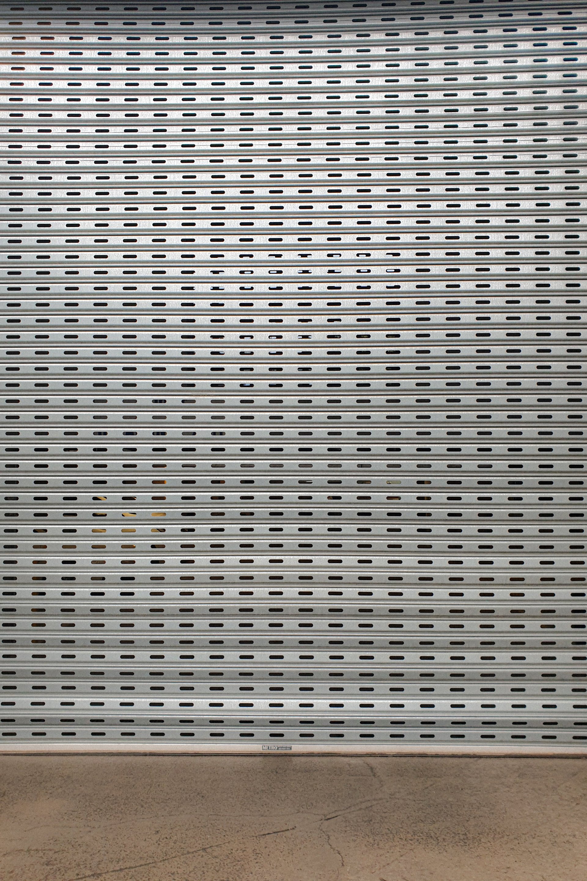 photo of a silver, perforated roller shutter with a small area of brown polished concrete visible below it